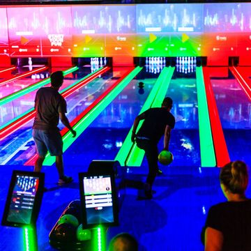 Bunt beleuchtete Bowling Bahnen mit Menschen, die den Ball rollen im Vordergrund. Bowling Hersteller Alternative.